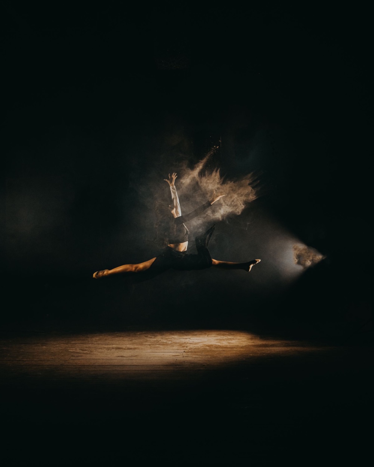 A dancer leaps in a full split through a single beam of stage light, a cloud of dust trailing from her hand.